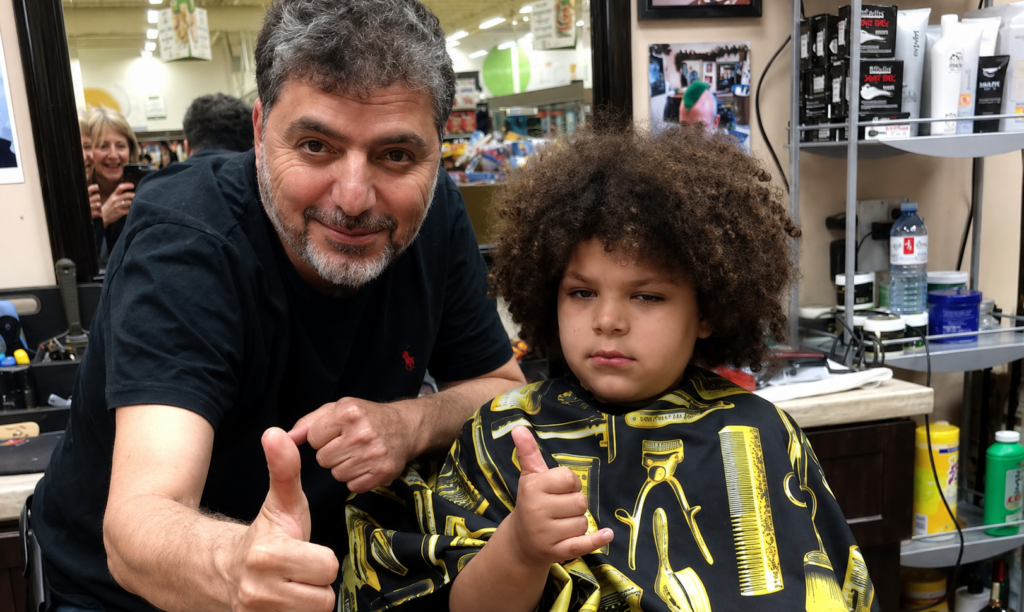 Ali, the owner of The Perfect Cut Barbershop, giving a thumbs up beside a young client in a barber cape after his haircut.